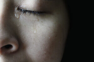 Close-up, monochromatic photo of a woman, just her nose, left eye, and cheek. Her eye is closed. At the inside corner, a tear has formed. In the next instant it will start running down her cheek.