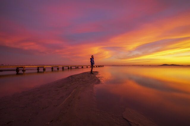 In this photo there's a figure of someone standing on a narrow spit of sand jutting into the water, a lake I'm guessing. This someone is looking up at the sky, which is filled with yellows, oranges, pinks, purples, and reds, which are then reflected in the water below. Is this a sunrise or sunset? I don't know. On the image sites you can sometimes find the same photo in both search categories.