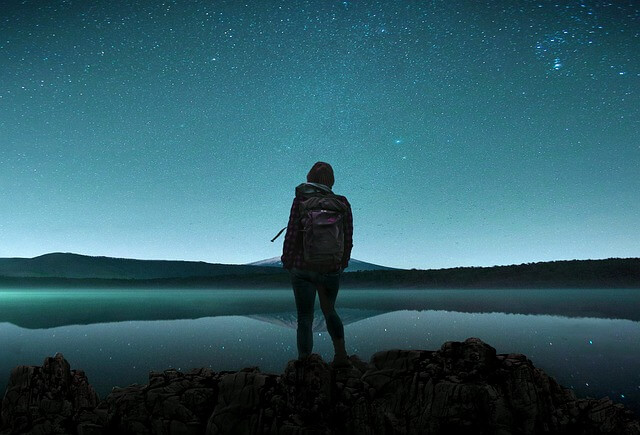 This is a photo that could be a painting. In the foreground is a camper with a hefty backpack. He or she is standing on a rocky outcropping. In mid-ground there is a lake and on the far side some gentle low hills. This is all done in black and green-blue. In the sky above night is falling. Behind the hills is a soft blue light turn to dark blue with a delicious hint of green undertones. You can see stars coming out to bless the camper's journey.