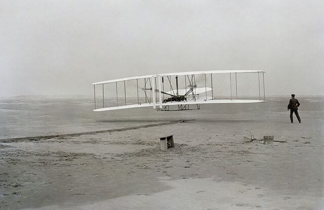 Iconic photo of the Wright brothers' first flight, one brother lying lying flat on his stomach between the upper and lower wings, the other brother standing off to the side attentive. Grey beach, grey sky.