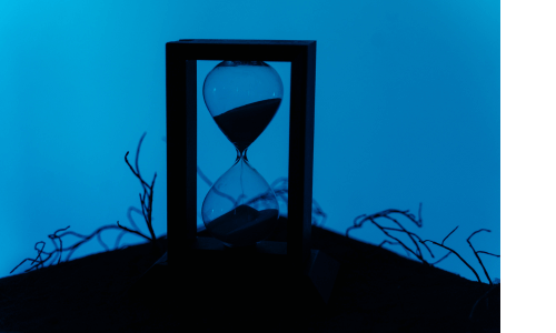 Hour-glass timer in a dark wooden frame sitting on a heap of black earth, a couple scraggly weeds flanking it. The background is a deep, rich blue, the exact color of sorrow.. The sand is running through.